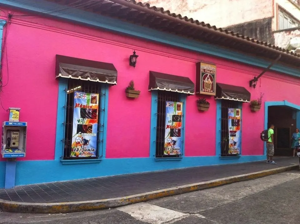 La fachada de la matriz en una etapa anterior: muro rosa con zoclo azul, toldos café y el letrero de El Itacate